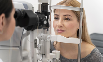Woman at an eye exam, seated at a slit lamp while an optometrist examines her eyes