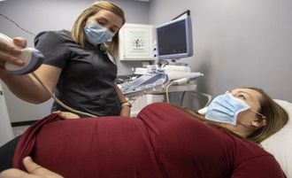 Dentist examining a patient in a dental clinic, both wearing masks, with equipment and monitor visible.