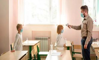 Teacher checks a student’s temperature in a bright classroom; two masked children sit at desks.