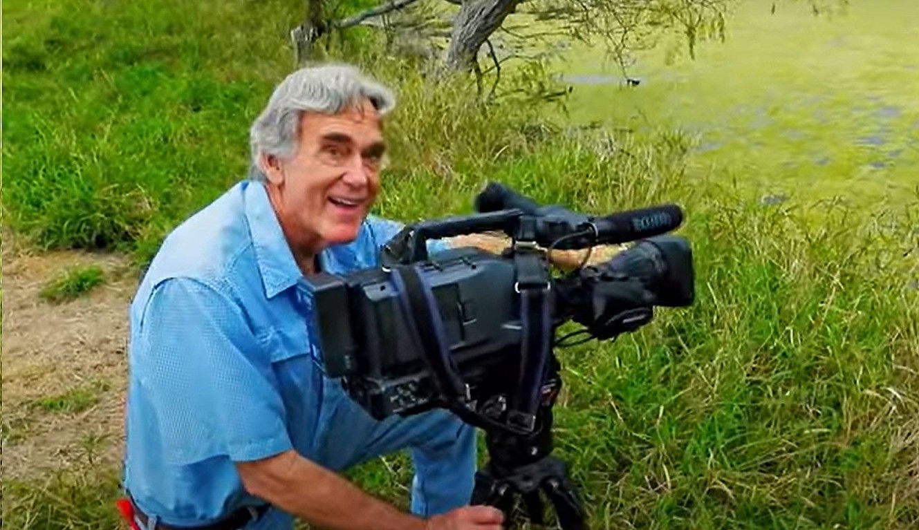 A man is kneeling down in front of a camera in a field.