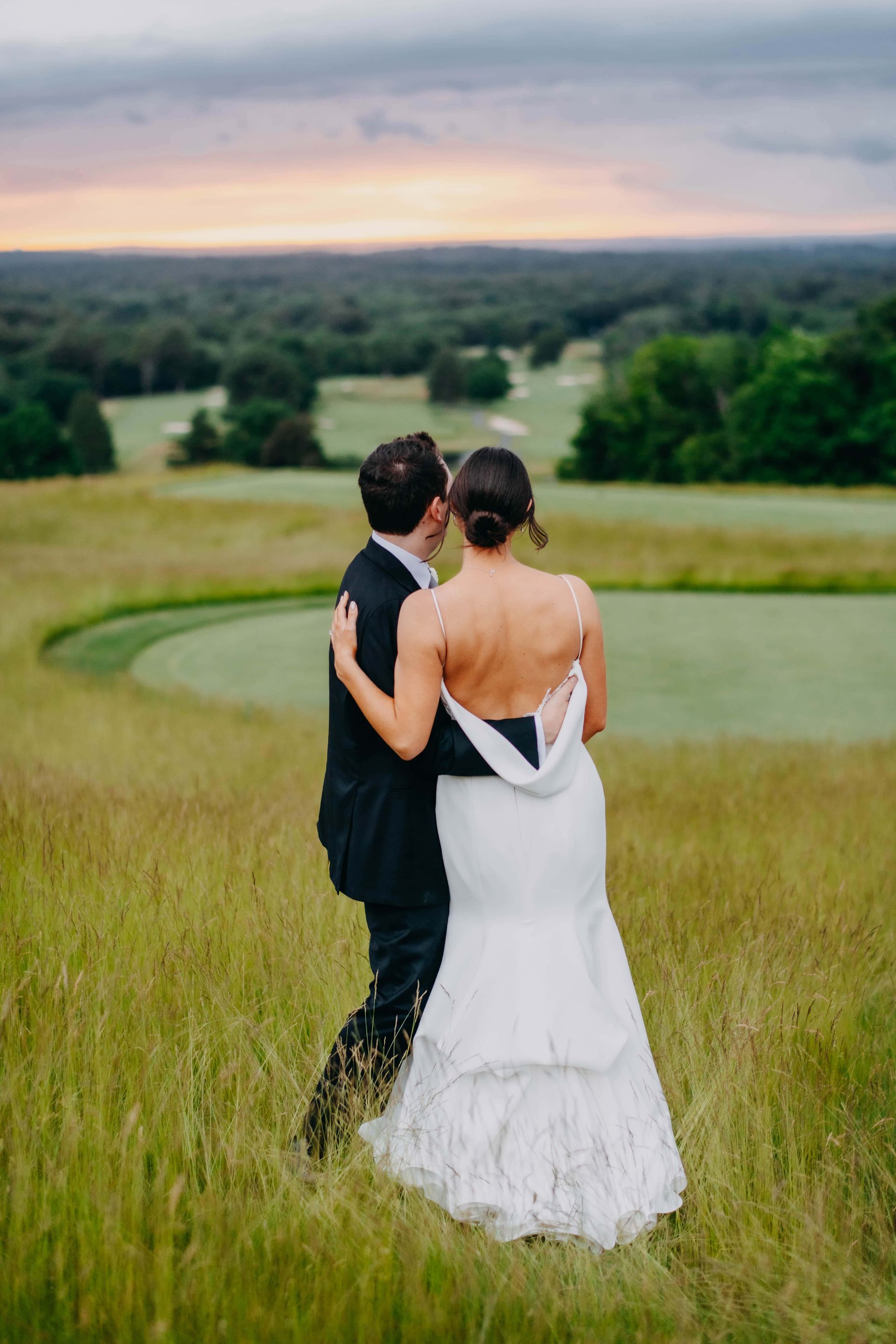 Bride and groom embrace on grassy hill overlooking The Starting Gate at GreatHorse.