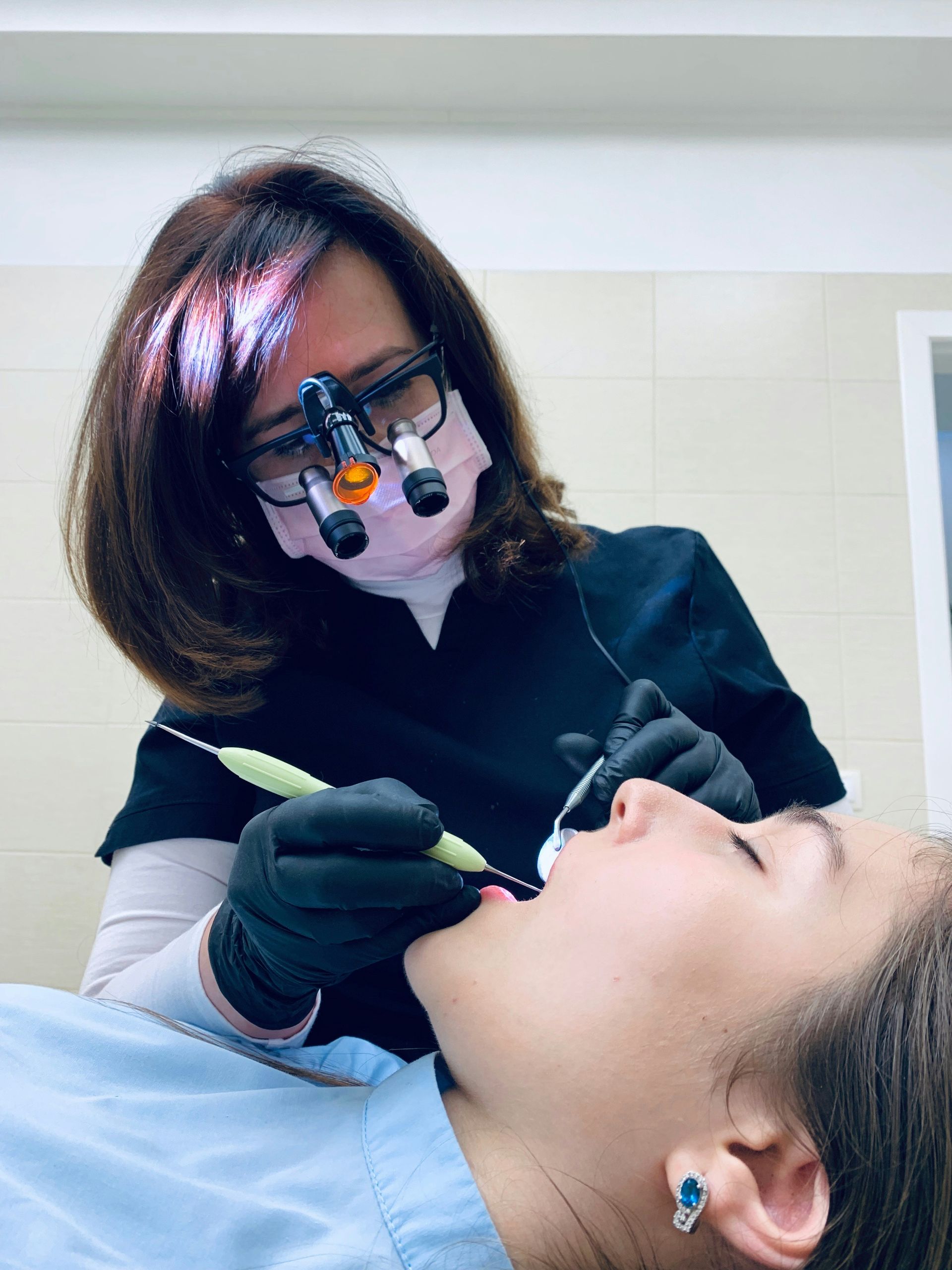 Dentist examining patient's teeth. She wears a mask, glasses, and loupes. The patient is in a dental chair.