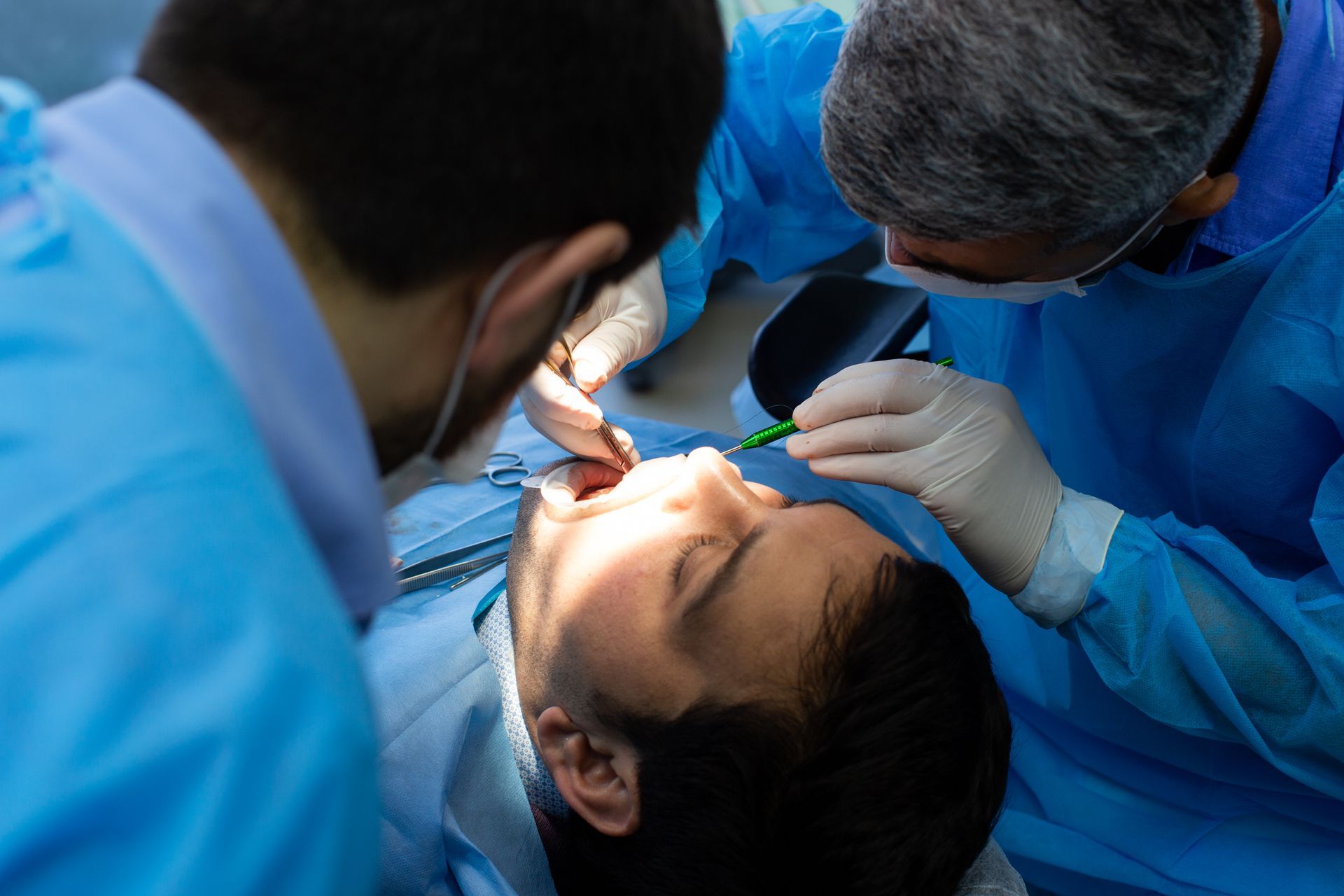 Two dentists in blue gowns examine a patient's teeth, using tools in a dental office.
