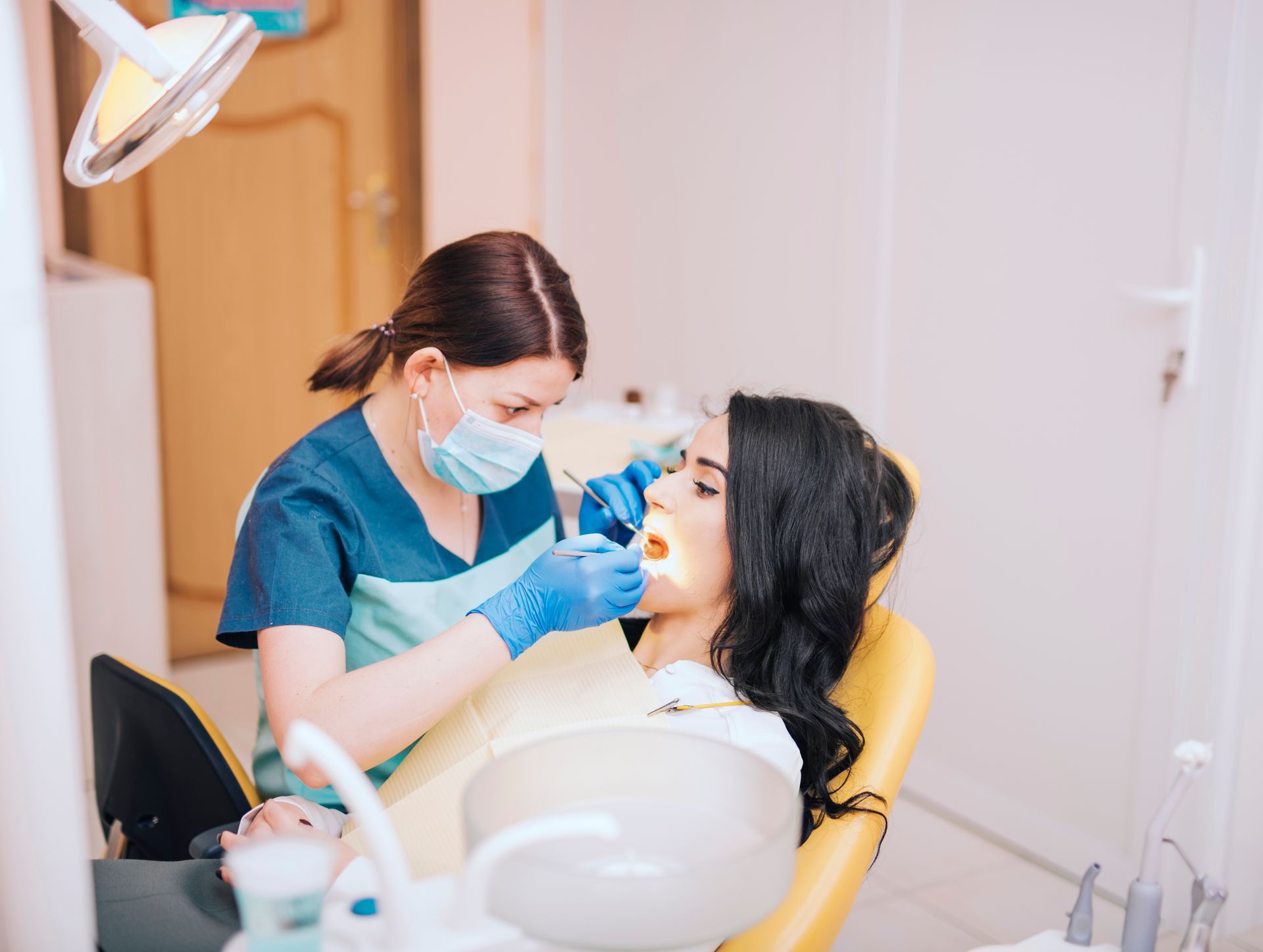 Dentist in blue scrubs examining patient’s teeth with tools. Patient reclined in yellow chair.