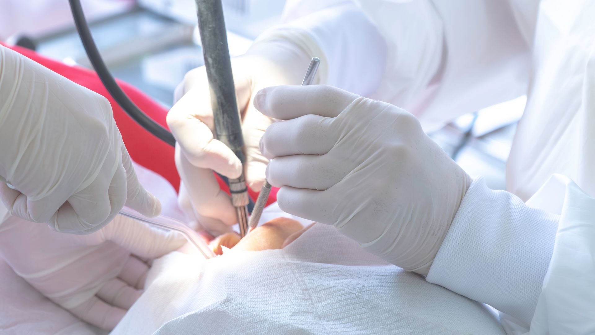 Dentist using tools on a patient's mouth, hands gloved. Red chair, white bib.