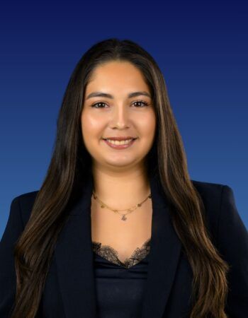 A professional headshot of a smiling person with long dark hair, wearing a dark blue blazer against a blue background.