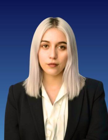 A person with shoulder-length blonde hair wearing a white collared shirt and black blazer against a blue background.