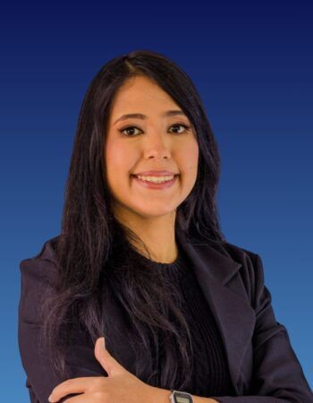 A smiling person with long dark hair, wearing a dark blazer over a black top, poses against a blue gradient background.