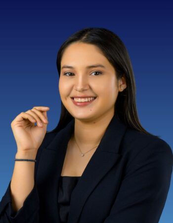A smiling individual in a dark blue blazer and simple necklace poses against a blue gradient background.