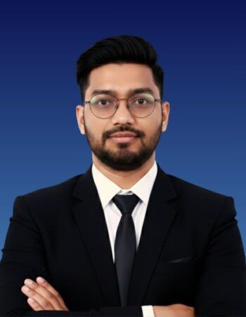 A man in a suit and tie with glasses, posing with arms crossed against a plain blue background.