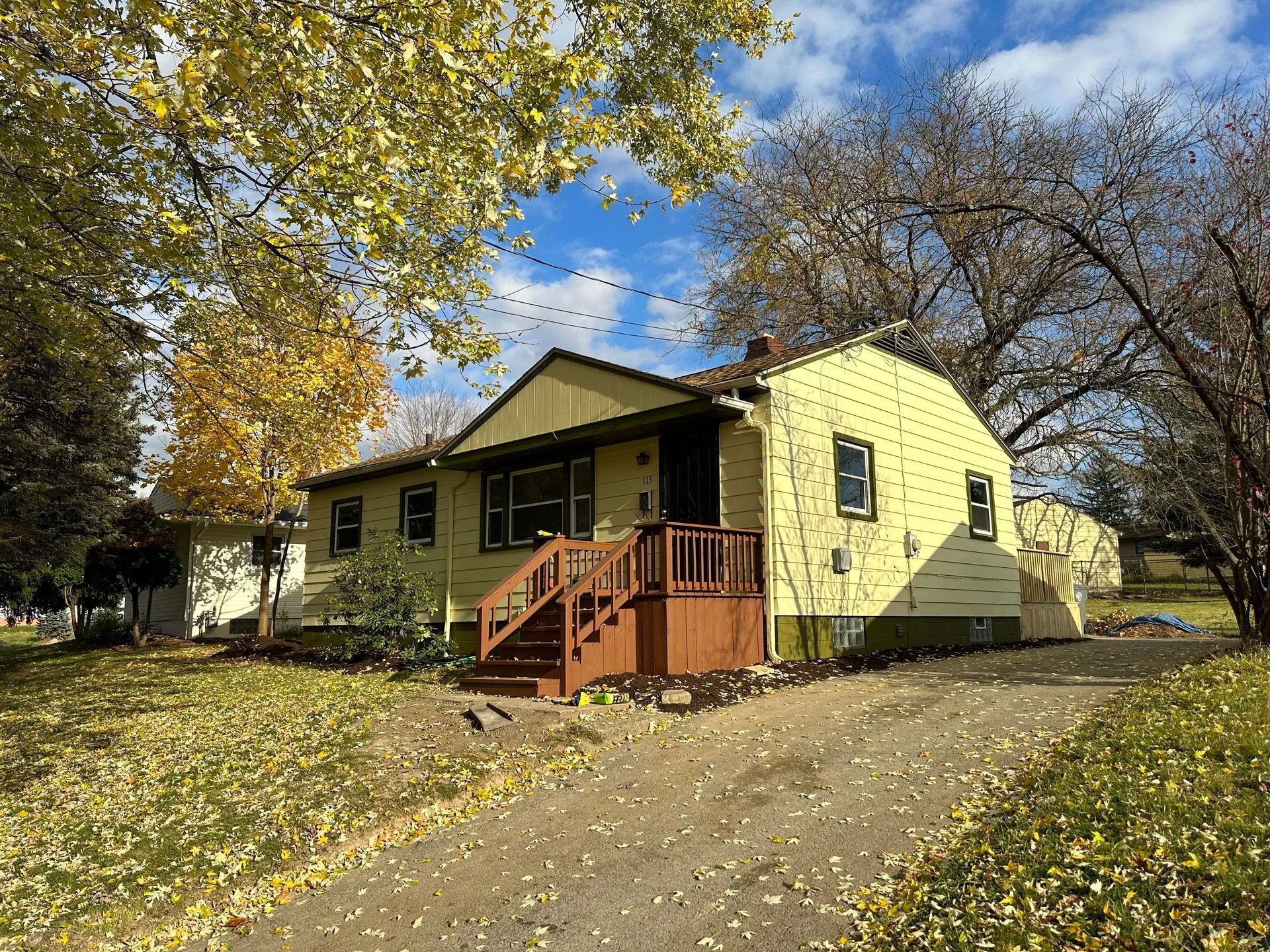 A yellow house with a porch and stairs is surrounded by trees and leaves.