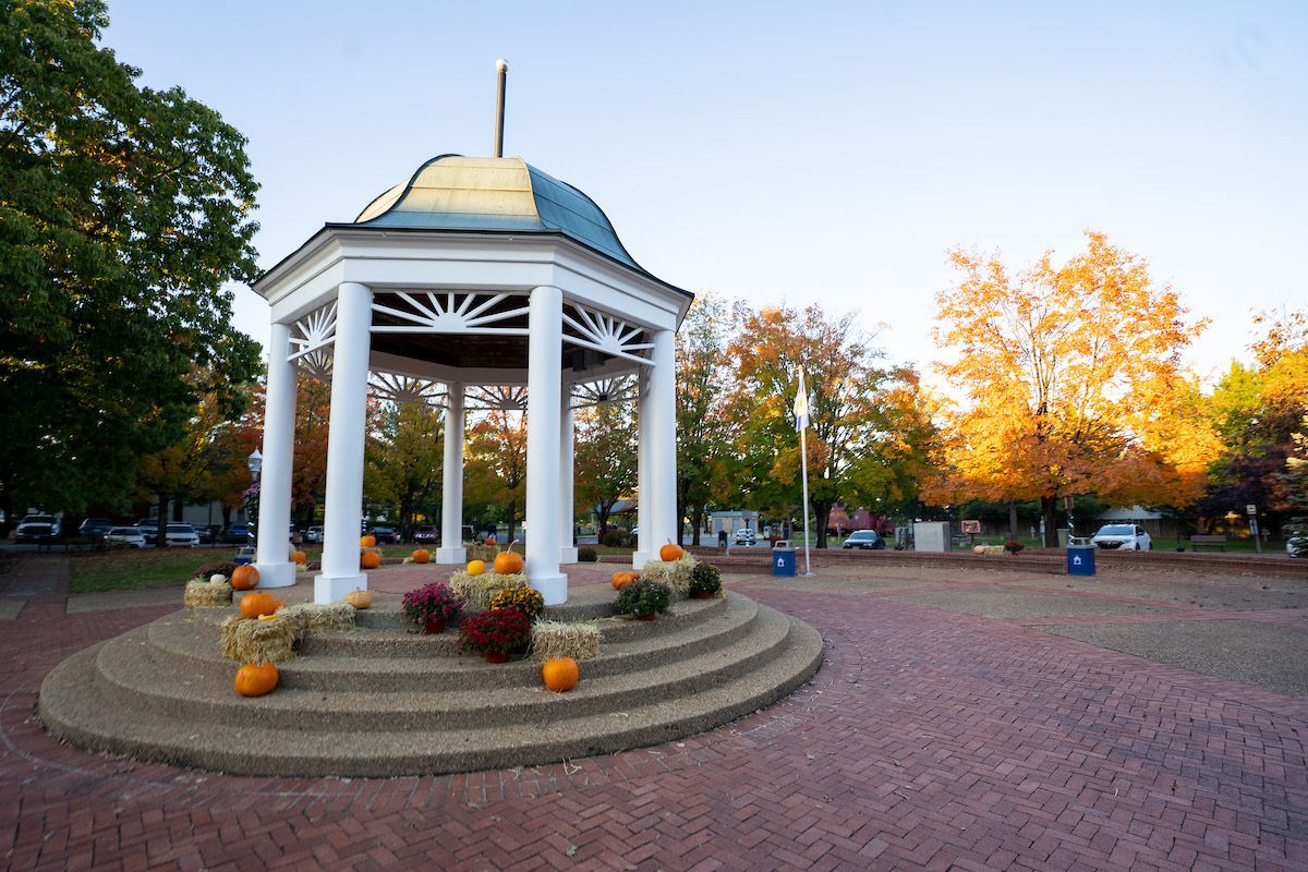 White gazebo with autumn pumpkins and flowers in a brick-paved park at sunset