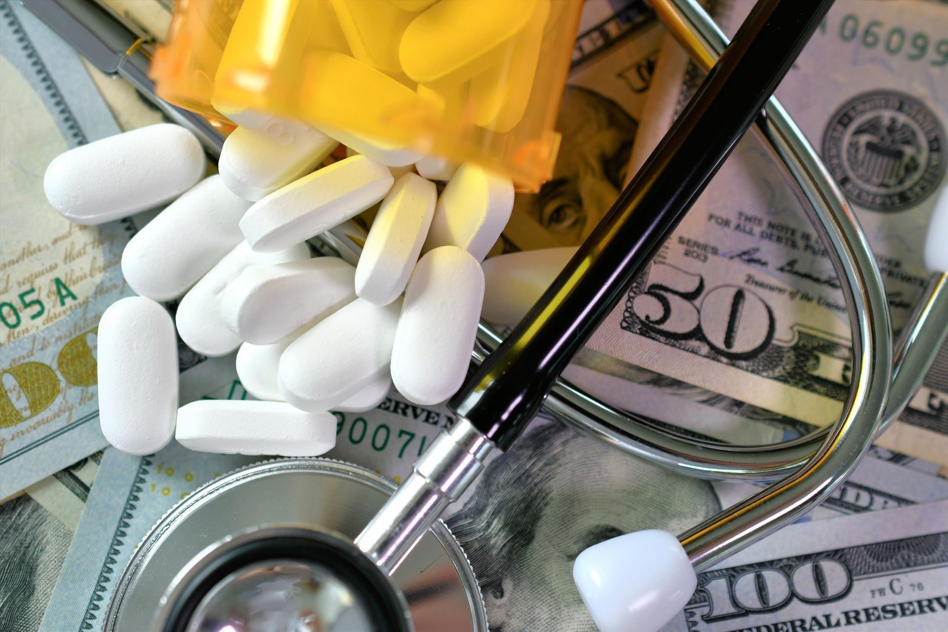 A stethoscope lies atop a pile of US currency bills with an open bottle of white pills spilled out.