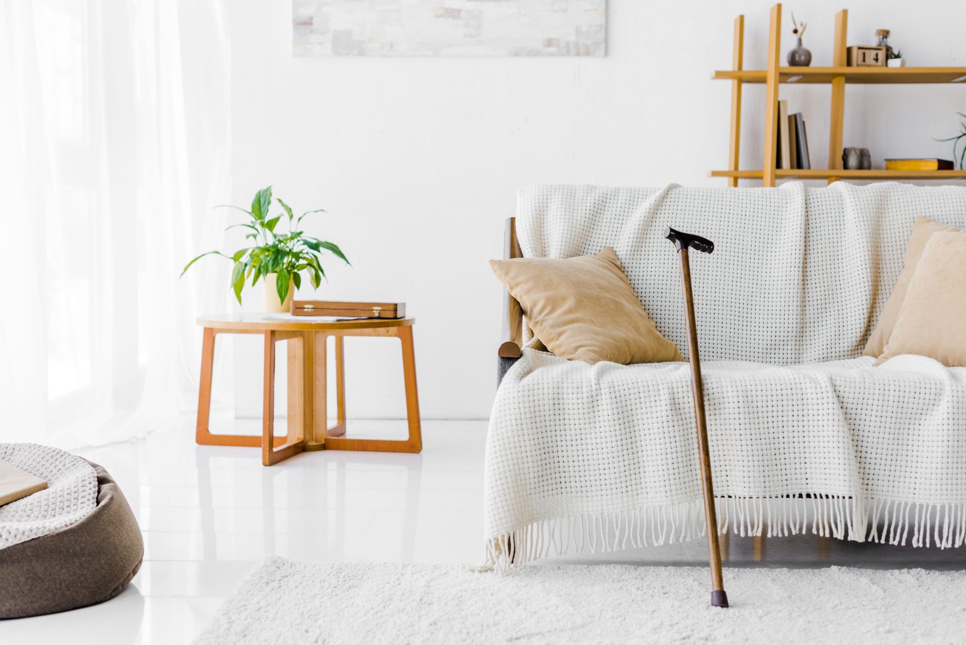A wooden cane stands upright against a couch with a beige pillow and white throw blanket in a bright living room.