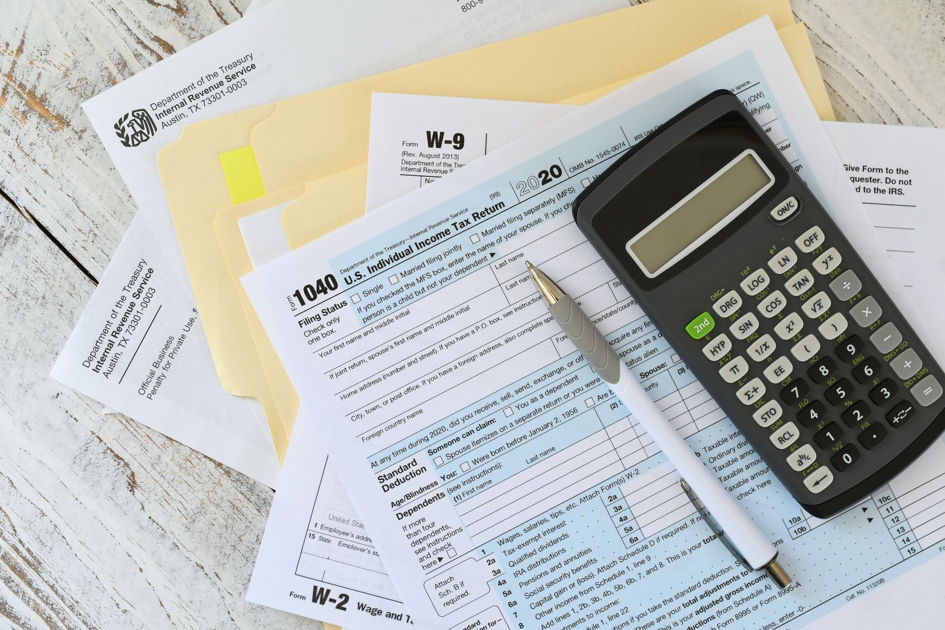 U.S. federal income tax forms, including a 1040, a calculator, and a pen, resting on a white wooden surface.