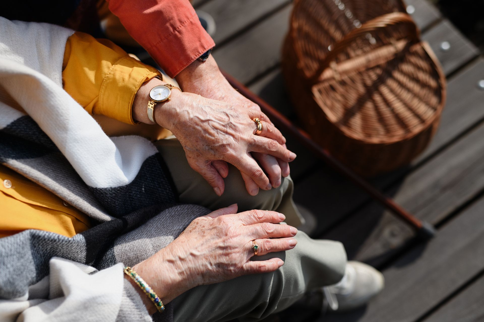 Two pairs of hands resting together on a lap, with a woven basket and wooden floor in the background.
