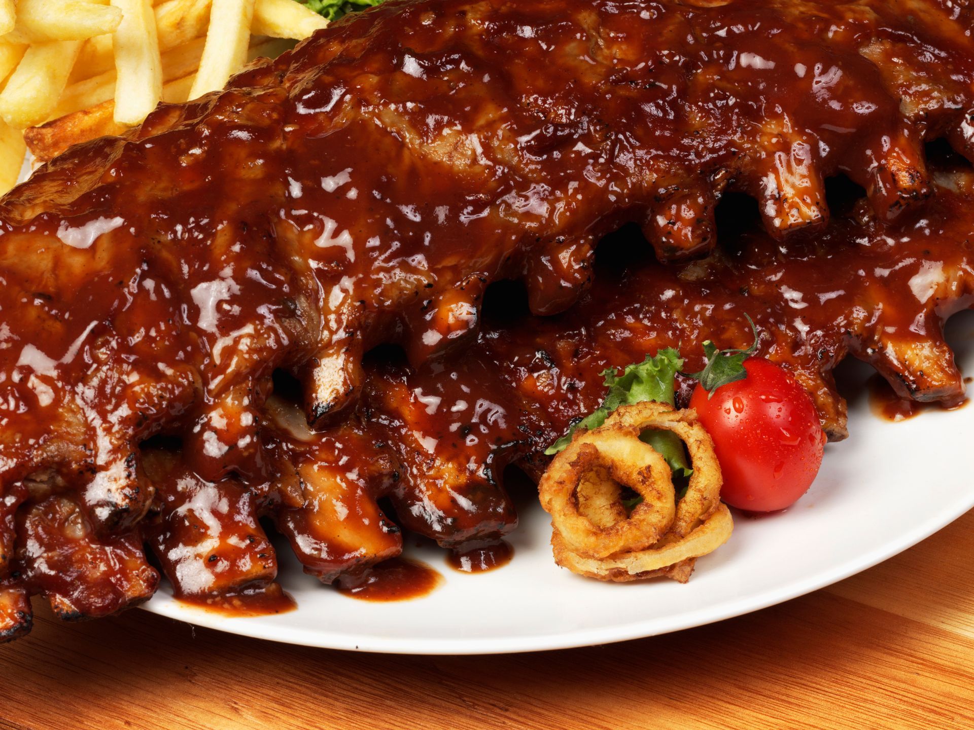 Rack of barbecued ribs with glossy sauce on a white plate, served with fries, onion rings, and a tomato.