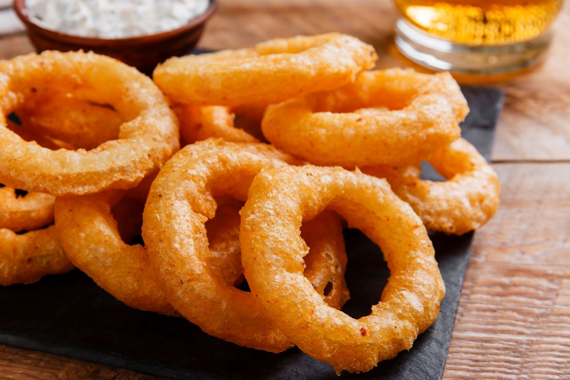 Golden-brown onion rings piled on a slate board, with a side of white dipping sauce and a glass of beer.