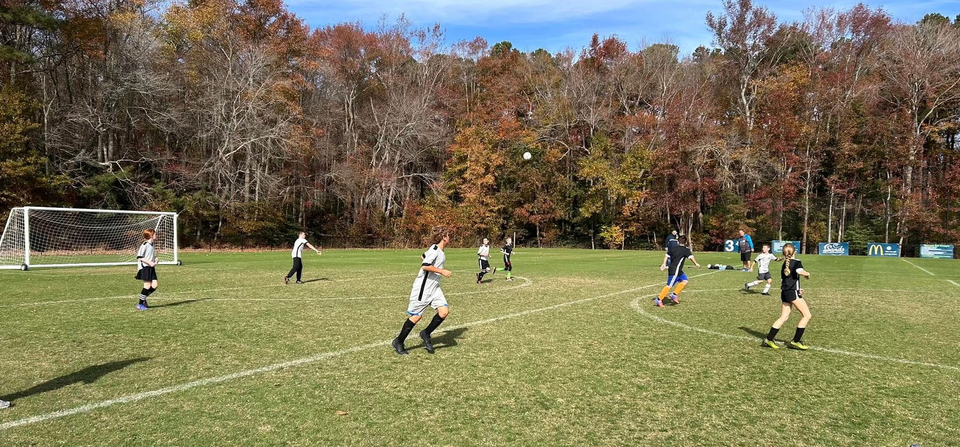 Soccer game in progress on a grassy field, with players in black and white uniforms, and autumn trees in the background.