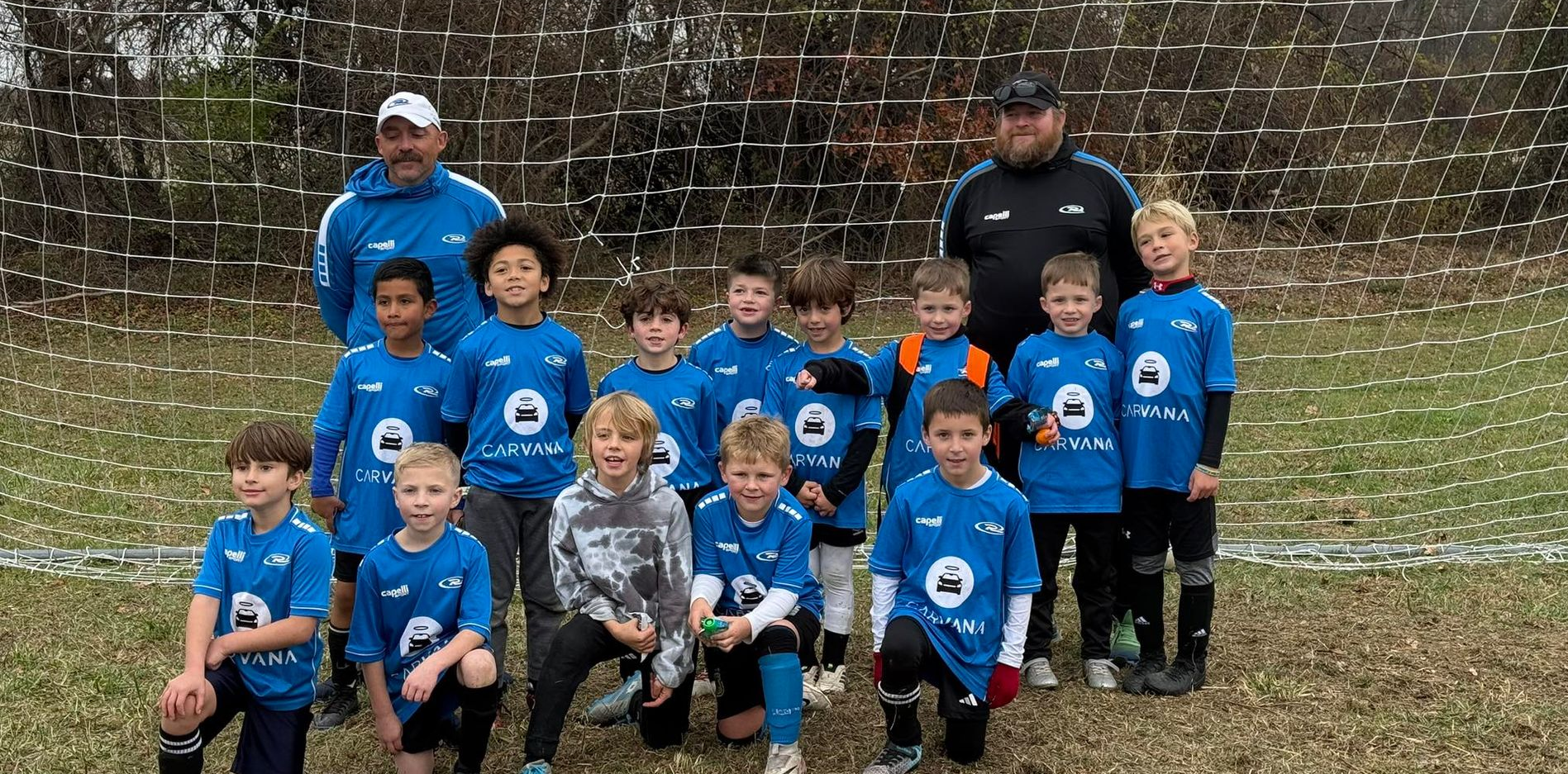 Soccer team of children and coaches posing on a grassy field. They wear blue shirts with a white logo.