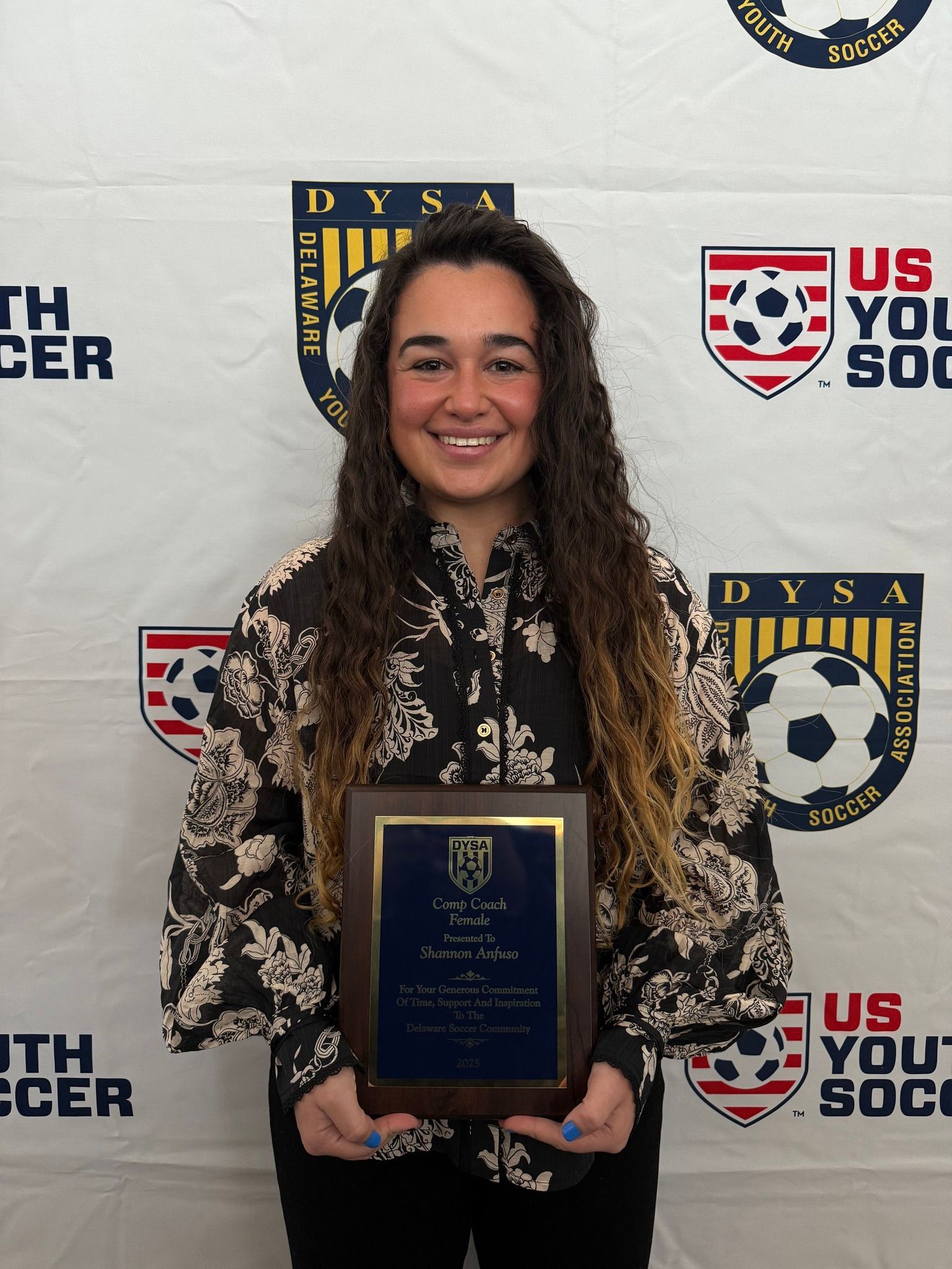 Woman holding a plaque, smiling, in front of a US Youth Soccer backdrop.