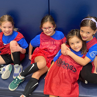 Four young girls in red and blue sports uniforms, smiling and posing indoors.