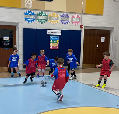 Children playing soccer in a gym. Some wear red vests; others, blue shirts. Light blue court, banners on wall.