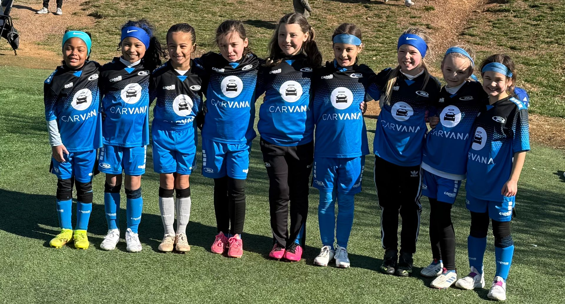 A youth soccer team in blue and black uniforms stands in a line on a grassy field, posing for a group photo.