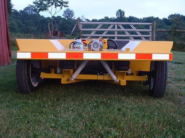 Rear view of a yellow trailer with reflective safety strips, tires, and mechanical components, on a grassy field.