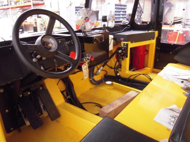 Interior view of a yellow industrial vehicle cabin with a steering wheel, pedals, and black control panel.