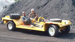 Two people in yellow open-top vehicle, spare tires, coal pile in background.