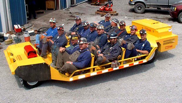 Group of miners in a yellow vehicle. All wearing helmets, driving outdoors.