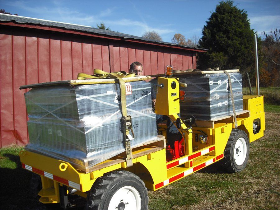 Yellow industrial vehicle carrying two wrapped pallets outdoors; a person is visible.