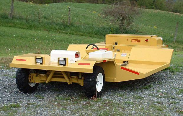 Yellow heavy-duty mower vehicle on gravel, grassy field background.