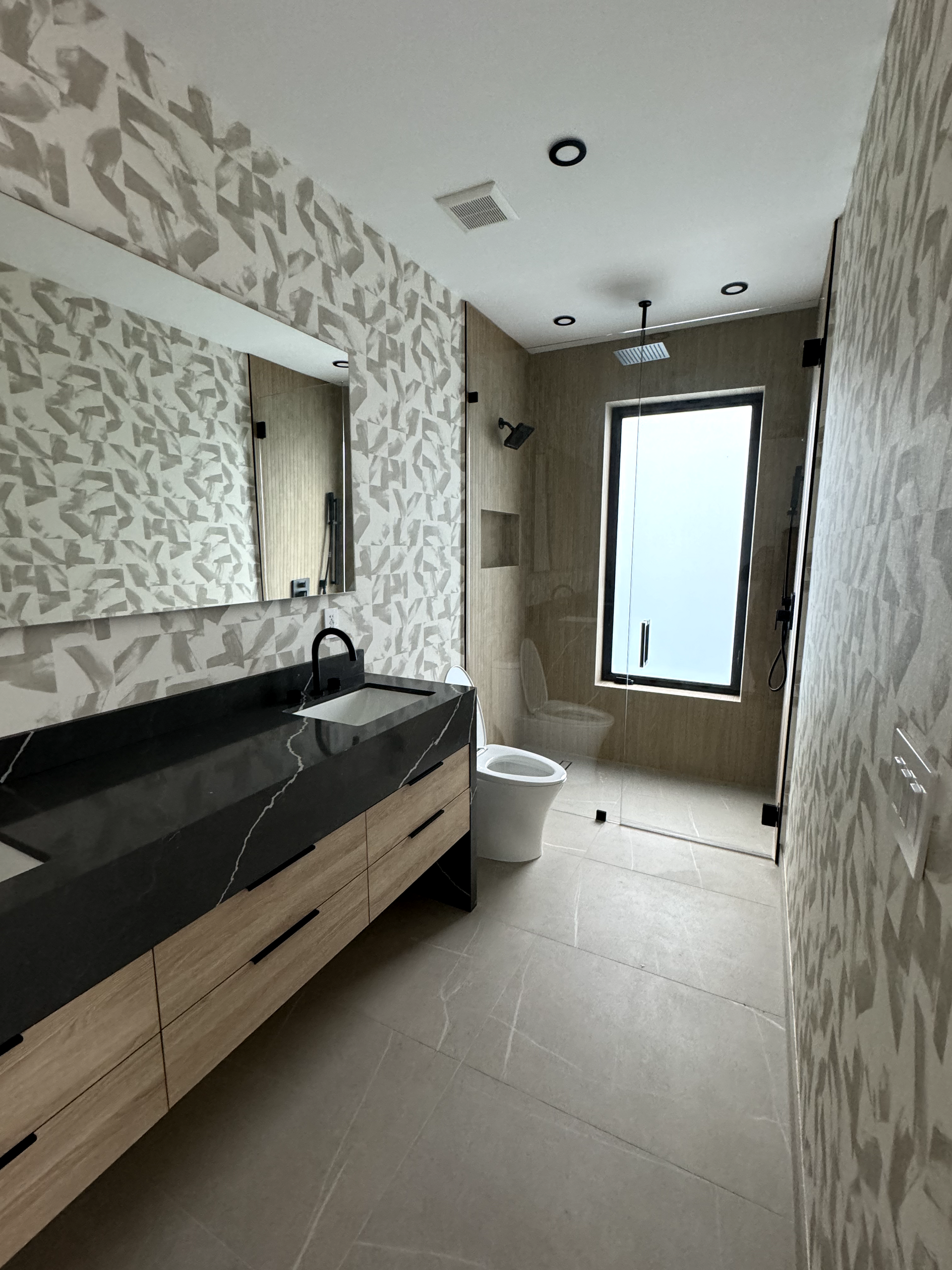 Modern bathroom with black countertop, wood cabinets, patterned wallpaper, and glass shower.