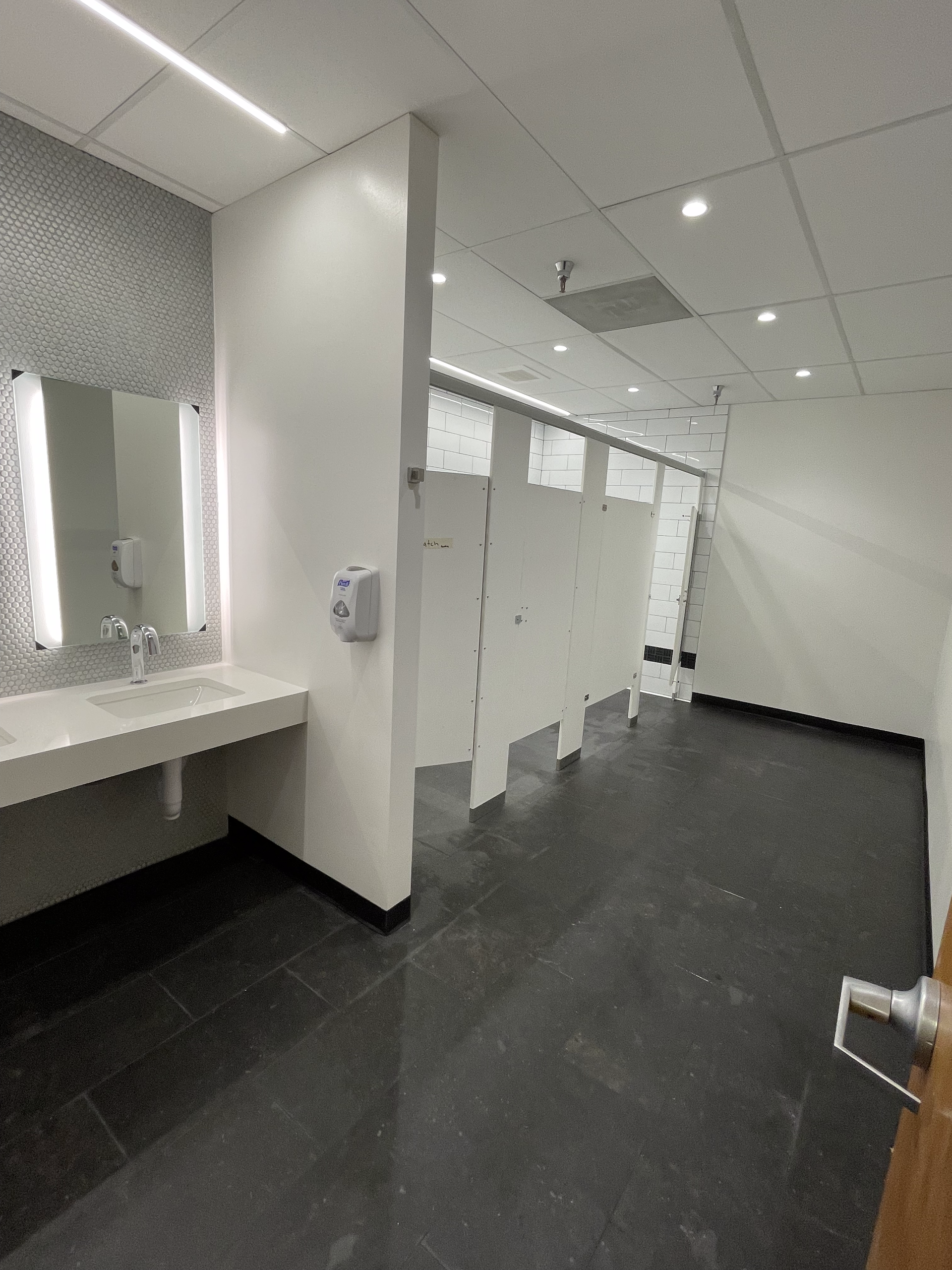 Bathroom interior with white stalls, a long sink with a mirror, and dark flooring.