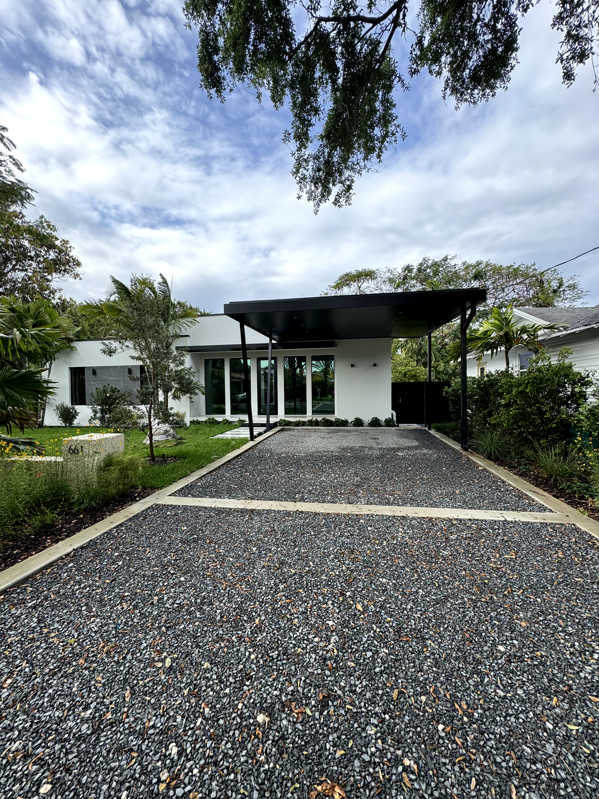 White house with dark carport, gravel driveway, and surrounding greenery under a cloudy sky.