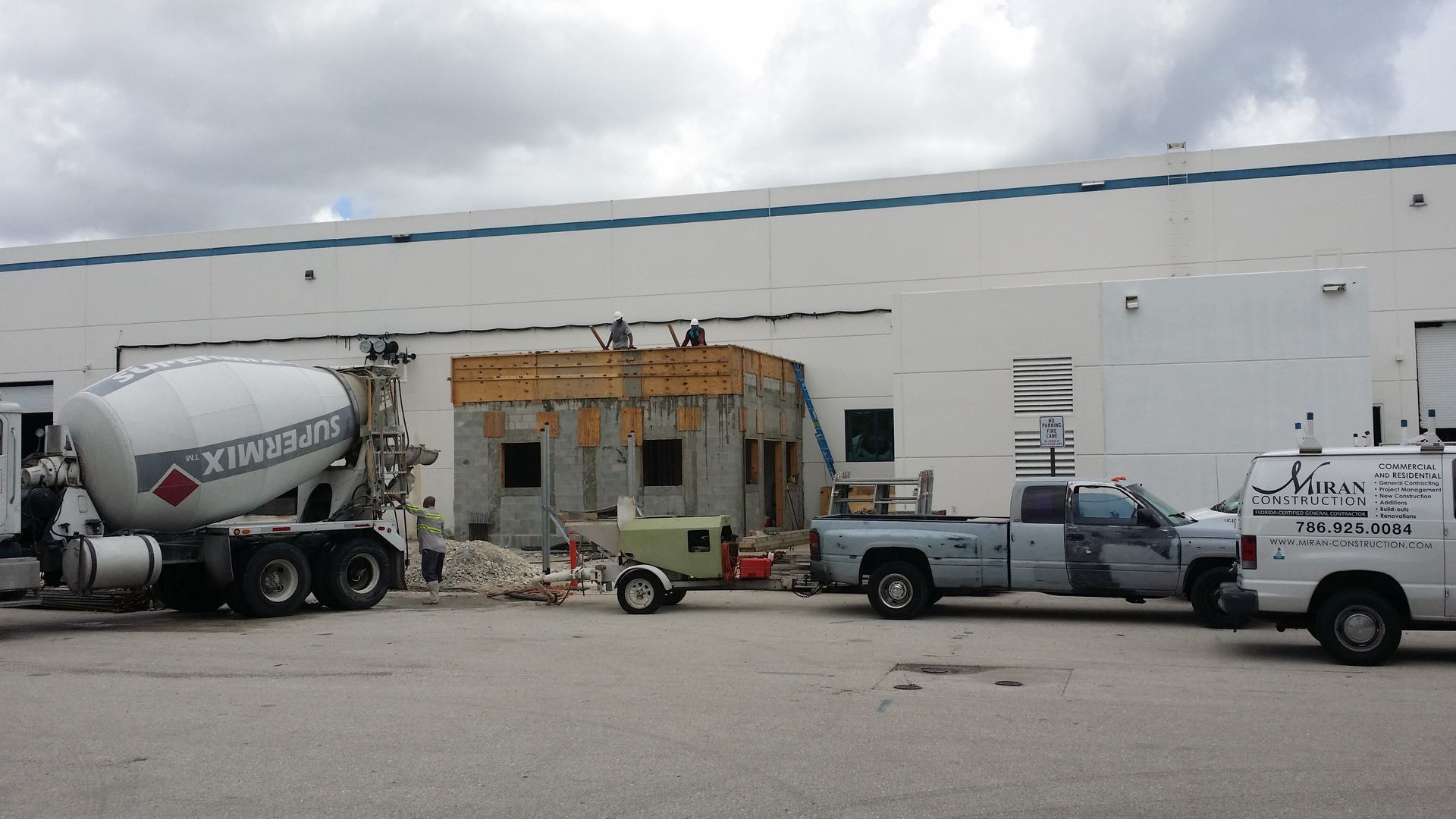 Construction site with a concrete mixer truck, workers, and trucks by a building.