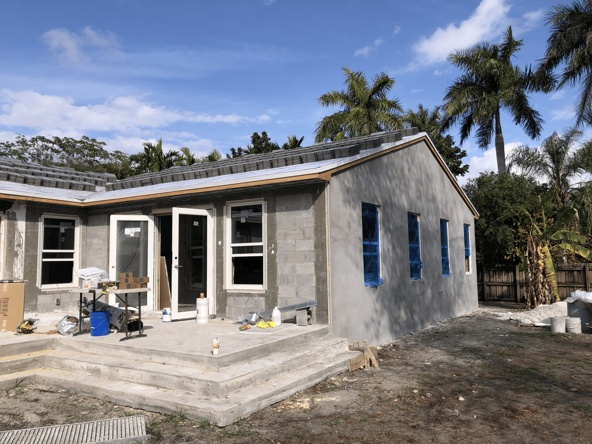House under construction: concrete block walls, windows/doors, stucco finish, steps, partially roofed, blue sky.