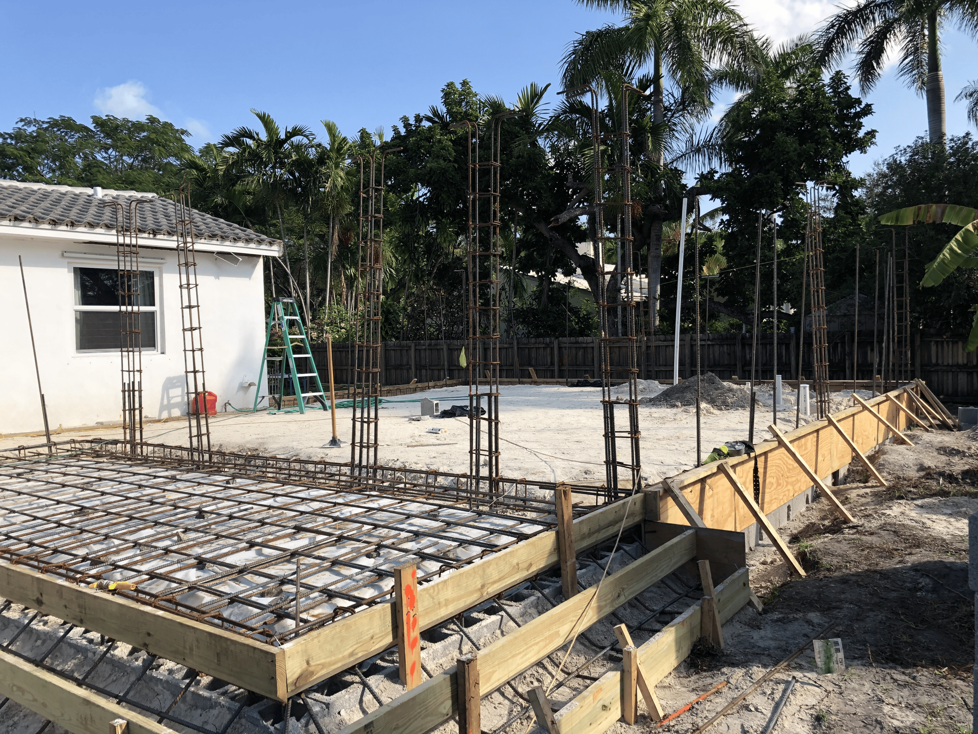 Construction site with wooden forms, rebar framework, and a partially built foundation in front of a white building.