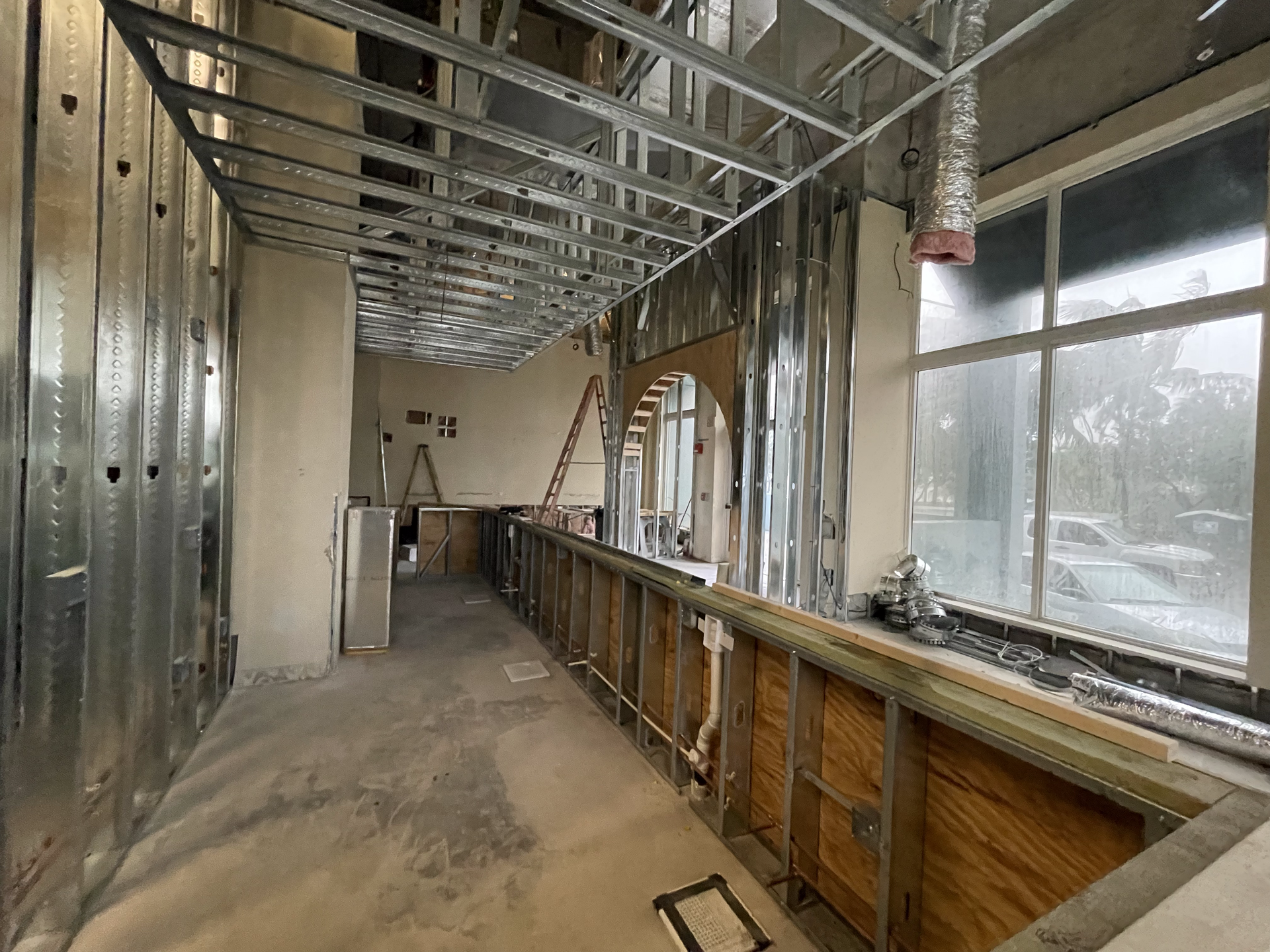Interior construction site with metal studs, unfinished walls, and windows looking out at trees.