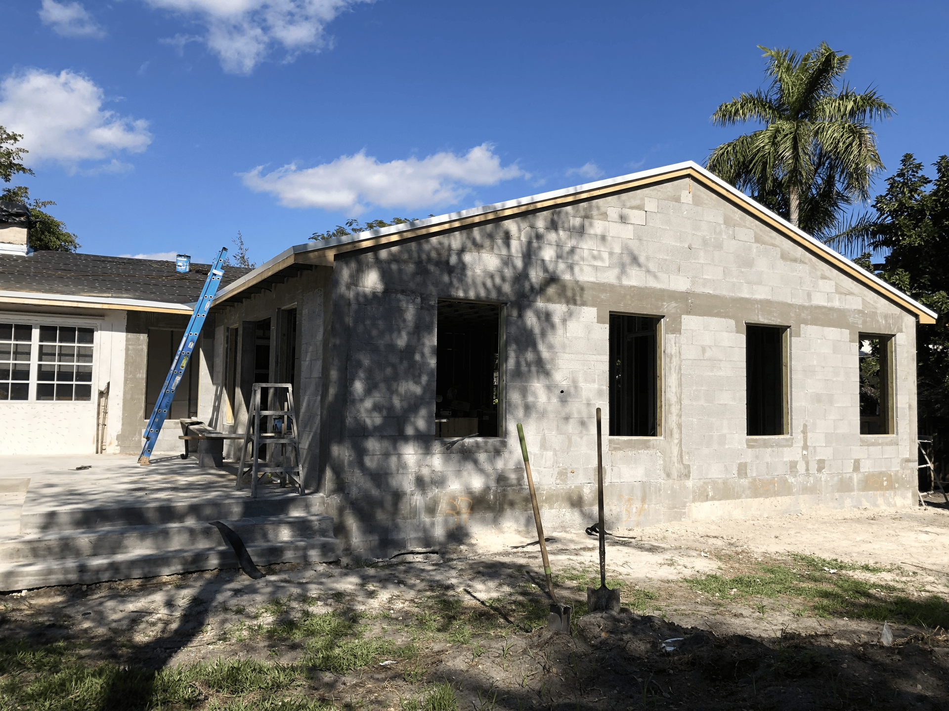 Construction of a concrete block building under a blue sky, with unfinished windows and a ladder.