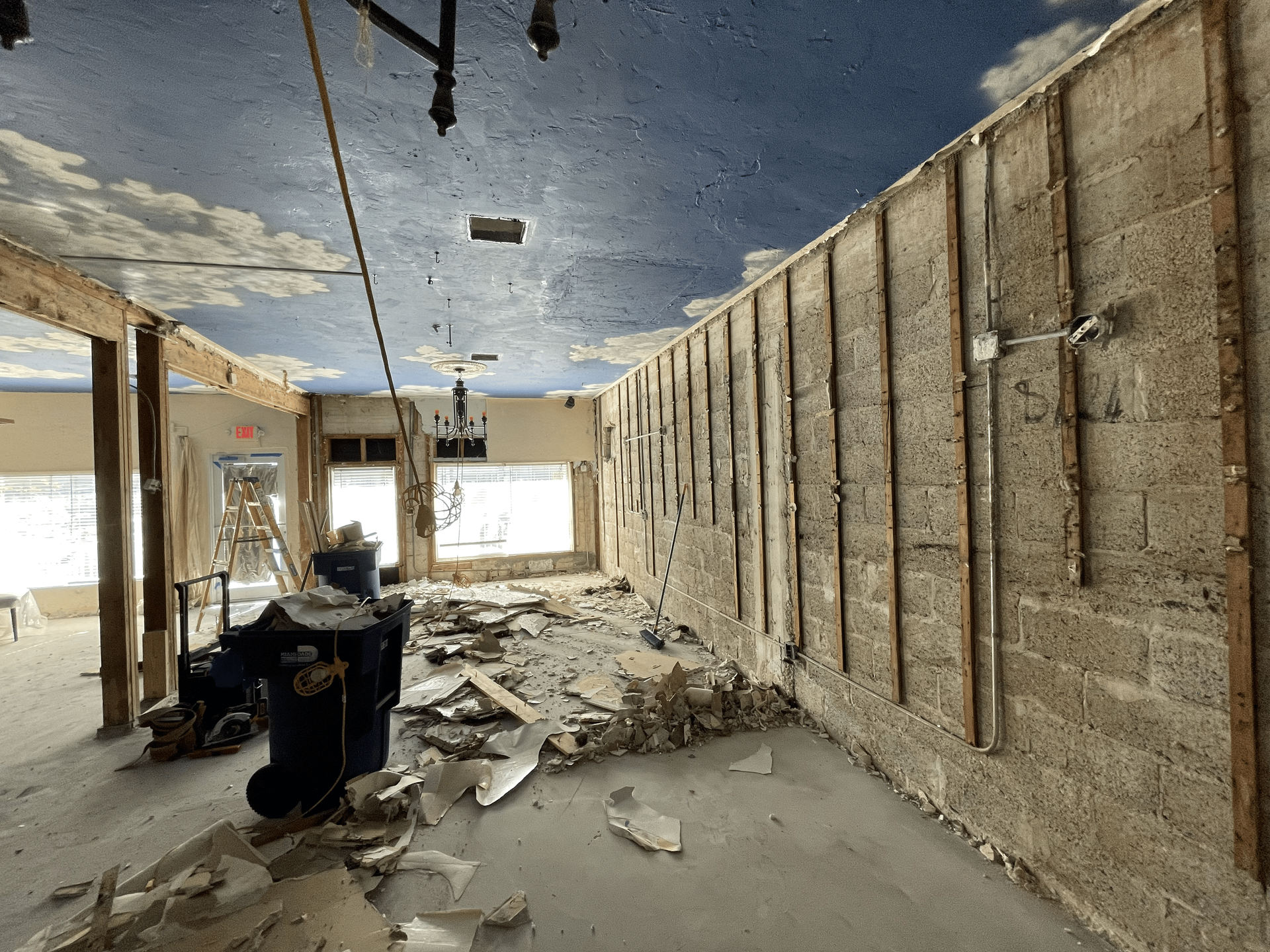 Interior demolition: walls stripped, debris on floor, sky-painted ceiling, and a worker.