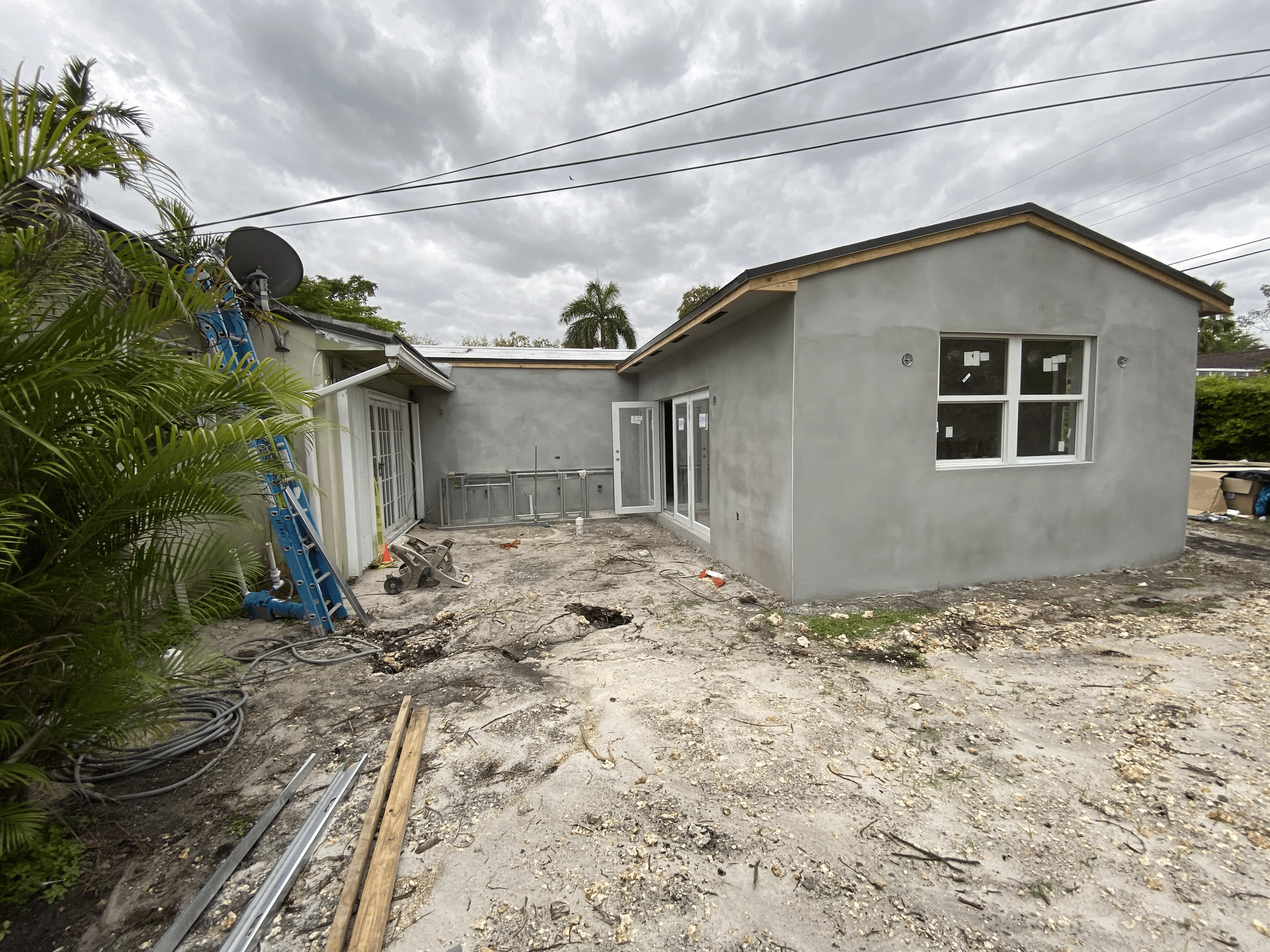 Exterior of a house under construction. Gray stucco walls, new windows and doors, debris-filled yard, overcast sky.