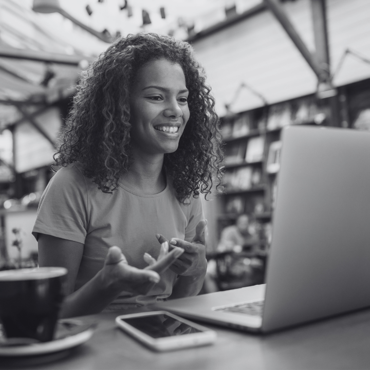 Woman smiles while on a video call, gesturing with hands, at a cafe.