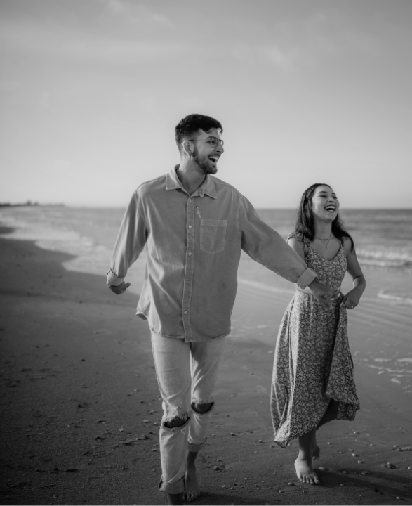 Couple running and laughing on a beach. Man in light shirt and jeans, woman in floral dress. Black and white.