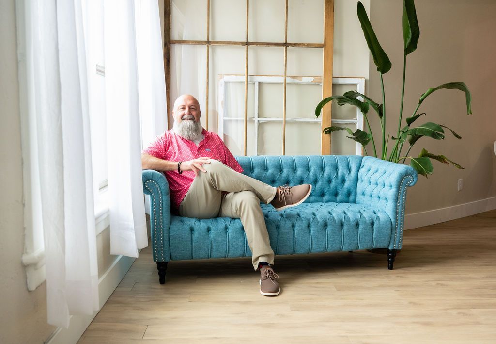 Man with a beard smiles, sitting on a blue tufted sofa. He wears a pink shirt and khaki pants.