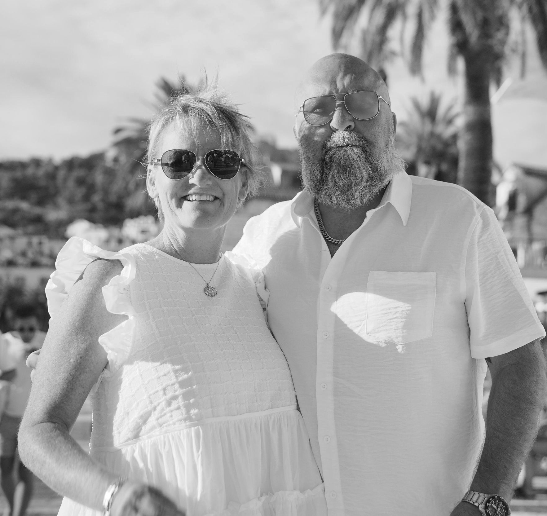 Couple in white clothing, smiling, posing outdoors with palm trees in background.