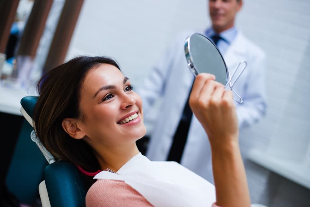 A Woman is Sitting in a Dental Chair Looking at Her Teeth in a Mirror — Advanced Dental Southern Highlands in Moss Vale, NSW