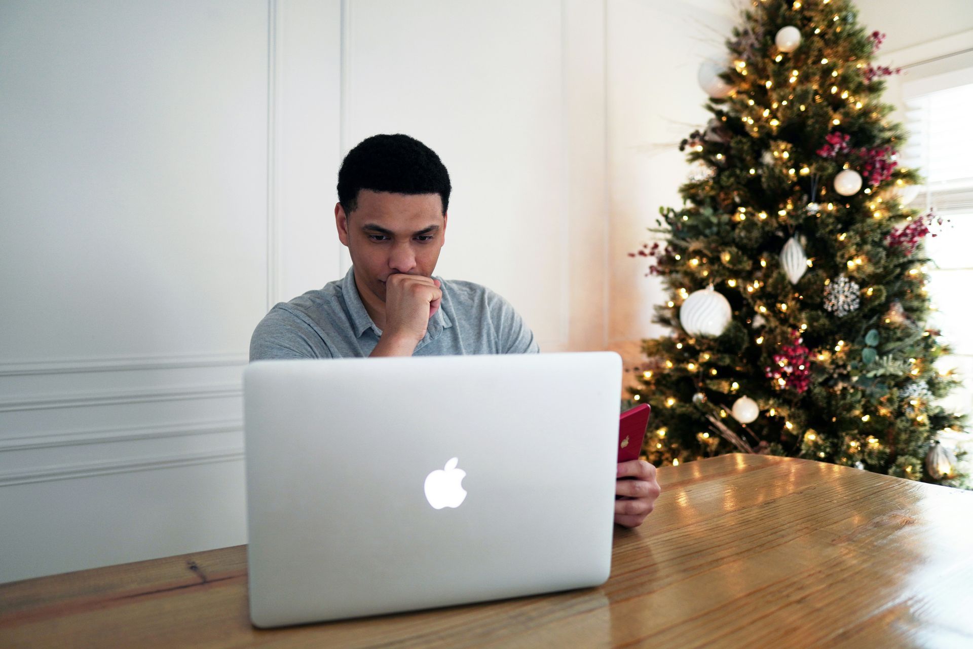 Man sitting at table, looking at laptop, with Christmas tree in background